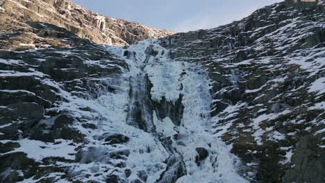 Frozen-waterfall-cascading-down-rugged-cliffs-in-Norway,-sunlight-glancing-off-the-icy-surface