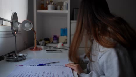 Side-close-up-of-young-girl-studying-on-desk-in-dark,-turns-on-lamp-close-to-her