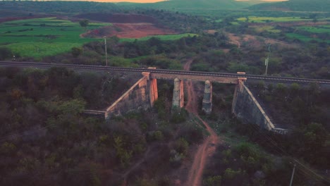 Luftaufnahme-Einer-Alten-Eisenbahnbrücke-Aus-Beton-Mit-Gleisen-Und-Dichten-Waldhügeln-Im-Hintergrund-Während-Der-Späten-Abendzeit-Per-Drohne