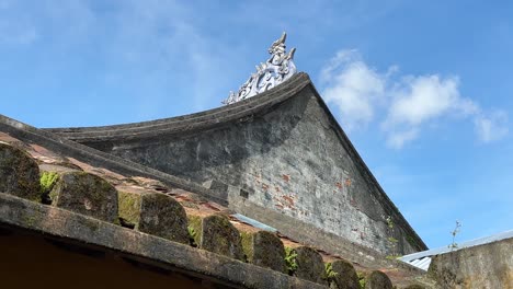 Temple-pitched-roof-with-cultural-centrepiece-on-apex-and-moss-in-Hoi-An,-Vietnam-ancient-town-with-blue-sky-background