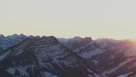 Witness-the-enchanting-sunrise-casting-a-golden-glow-on-the-silhouette-of-a-snow-covered-mountain-range,-captured-by-drone