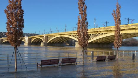 Flooded-benches-and-trees-at-the-quay-in-Budapest-with-Margaret-Bridge-in-the-background-during-the-flooding-of-Danube-River,-Hungary---December-28,-2023