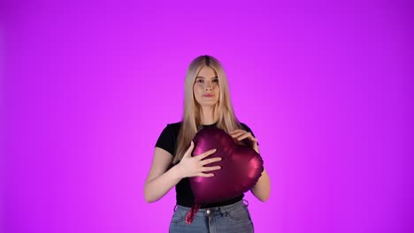 Cute-woman-looking-at-camera-and-holds-red-balloon-heart,-purple-background