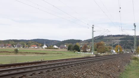 Red-Deutsche-Bahn-train-approaching-on-tracks-through-a-rural-landscape-with-houses-and-church-in-the-background,-daytime