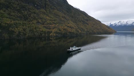 Autumn-colors-embrace-Geirangerfjord-as-a-ferry-crosses-tranquil-waters-with-snow-capped-peaks,-aerial-view