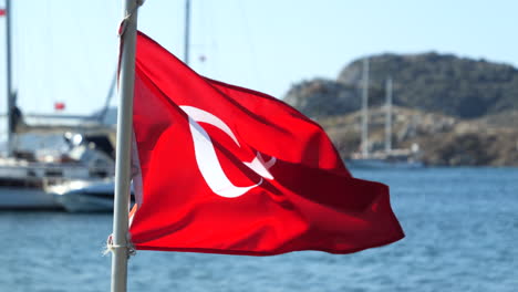 Bandera-Turca-Ondeando-Con-El-Viento-Con-Barco-Y-Mar-Al-Fondo-En-Gumusluk,-Turquía