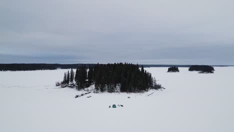 Eine-Drohne-Mit-Weitem-Landschaftsfoto-Einer-Insel-Auf-Dem-Zugefrorenen-Kanadischen-Paint-Lake-Mit-Einer-Eisfischerhütte-Und-Skioos