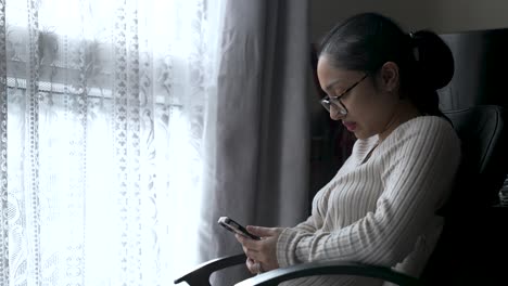 Mujer-Con-Suéter-Usando-Un-Teléfono-Inteligente-Junto-A-La-Ventana,-Luz-Natural,-Ambiente-Sereno