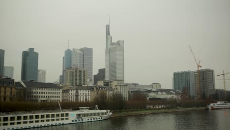 Overcast-day-view-of-Frankfurt-skyline-with-river-and-boats,-cityscape,-construction-cranes-in-background