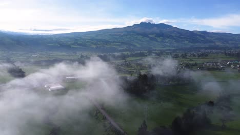 Slow-moving-drone-clip-over-a-green-field-and-a-village-with-the-volcano-Volcan-Rumiñahui-at-the-background-in-Neblina,-Machachi-in-Equador