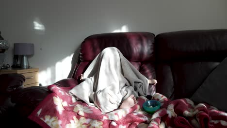 Funny-British-Asian-Toddler-Pulling-Blanket-Over-Himself-Sitting-On-Sofa-In-Living-Room-With-Morning-Sunlight-Coming-Through-Window