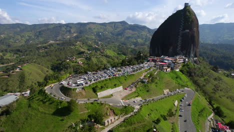 Guatape-Lake-and-El-Penol-Rock,-Aerial-View-of-Green-Landscape,-Road,-Parking-and-Landmark-on-Sunny-Day,-Revealing-Drone-Shot