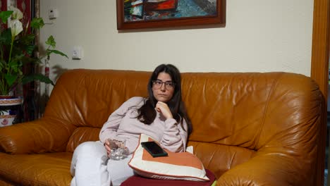 Woman-in-glasses-relaxing-on-a-brown-sofa-watching-TV,-taking-a-sip-from-a-glass