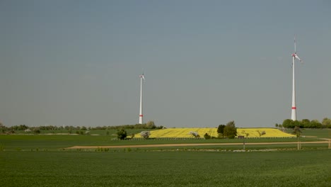 Wind-turbines-standing-tall-over-a-green-and-yellow-patchwork-field-under-a-clear-blue-sky,-static-shot
