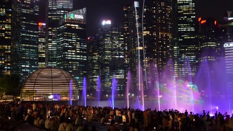Immersive-SPECTRA-light-and-water-show,-massive-crowd-of-audiences-at-the-Marina-Bay-Sands-event-plaza-with-downtown-cityscape-in-the-background,-time-lapse-shot-of-vibrant-night-of-Singapore-city