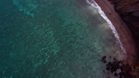 Stunning-aerial-view-of-tropical-Hawaiian-exotic-destination-with-turquoise-calm-ocean-surface,-coral-reef-and-red-dirt-cliffs