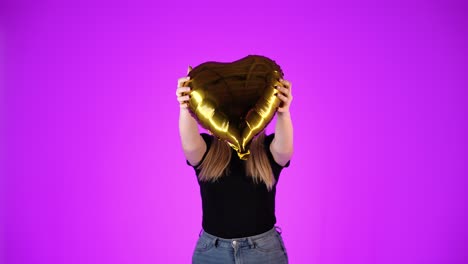 Woman-in-love-shows-golden-heart-and-smiles,-purple-background,-studio-shot