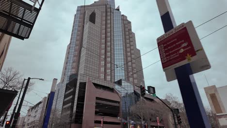 Moving-cinematic-footage-of-tall-corporate-building-in-sacramento,-california-with-a-grey-cloudy-sky-in-the-background-and-train-cables,-graffiti-and-street-signs-in-the-moving-foreground