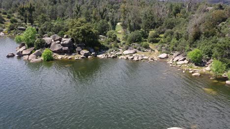 flight-in-retreat-and-ascent-in-a-reservoir-visualizing-the-shore-with-rocks-and-clear-water-and-discovering-a-forested-area-of-pines-and-fir-trees-on-a-spring-morning-in-Avila-Spain