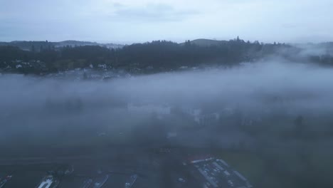 Drone-approaching-a-lakeside-village-shrouded-heavily-by-mist-and-fog,-located-in-the-county-of-Cumbria-in-United-Kingdom