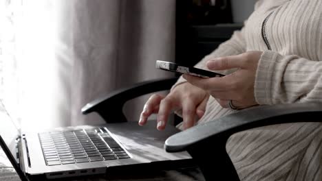 A-person-multitasks-by-typing-on-a-laptop-keyboard-while-holding-a-mobile-phone-in-one-hand,-highlighting-the-seamless-integration-of-digital-devices-into-daily-routines-at-home
