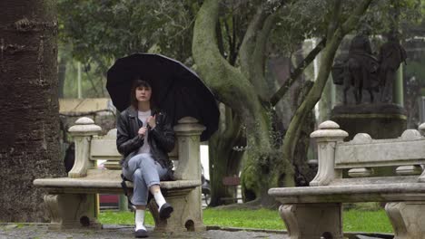 Traurige,-Frustrierte-Junge-Frau,-Die-Unter-Einem-Schwarzen-Regenschirm-Auf-Der-Bank-Im-Park-Sitzt