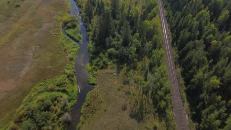 Tilt-Down-Aerial-Shot-Over-River-and-Railroad-with-Alpine-Forest-near-127-Mile-House,-Canada