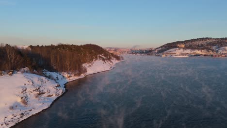 Luftaufnahmen-Von-Einer-Im-Winter-Schneebedeckten-Insel-Mit-Bäumen-Neben-Der-Wasserstraße-Mit-Schwebendem-Nebel-In-Norwegen