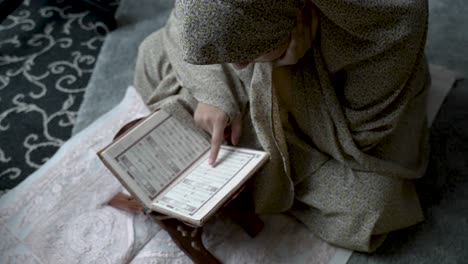 Seated-on-the-ground-amidst-stacked-clothing,-a-young-Muslim-girl-immerses-herself-in-a-religious-text,-embodying-the-notion-of-studying-and-acquiring-knowledge-in-the-cozy-confines-of-home