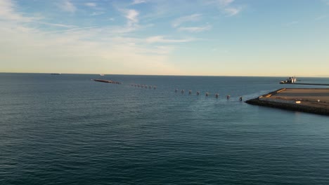 Vuelo-En-El-Mar-Viendo-Un-Antiguo-Muelle-Con-La-Pasarela-Casi-Desaparecida,-Solo-Quedan-Las-Anclas-Con-Barcos-Al-Fondo-Con-Un-Cielo-Azul-Con-Nubes-En-Un-Atardecer-De-Invierno-En-Valencia-España