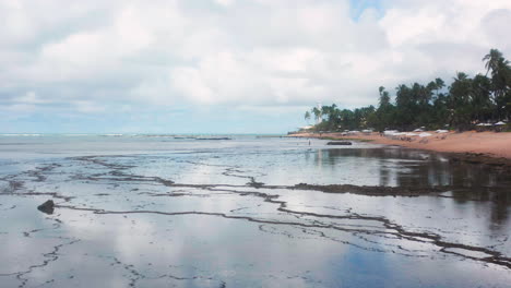 Vista-Aérea-De-La-Playa-Praia-Do-Forte,-El-Arrecife-De-Coral,-La-Zona-De-Palmeras-Y-Las-Nubes-Reflejándose-En-El-Océano,-Praia-Do-Forte,-Bahia,-Brasil