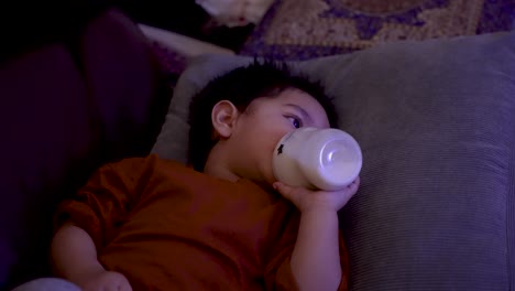 British-Asian-Toddler-in-orange-shirt-lying-down-drinking-from-bottle,-focused-on-TV-screen,-indoors