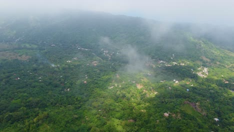 An-aerial-view-of-mist-covered-hills,-lush-greenery,-and-scattered-buildings-in-a-serene-landscape