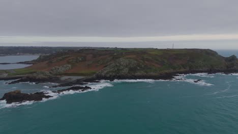 Guernsey-Pleinmont,-flight-from-out-to-sea-towards-the-south-west-tip-of-the-Island-on-cloudy-day-showing-cliffs,-beaches-and-rocks-and-the-BBC-communication-tower