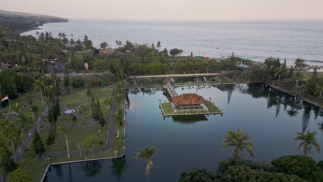 Eine-Ruhige-Luftaufnahme-Eines-Küstengebiets-Mit-Einem-Großen-Wassertempel-Taman-Ujung,-Umgeben-Von-üppigem-Grün-Und-Palmen,-Mit-Dem-Ruhigen-Meer-Im-Hintergrund