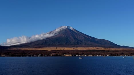 Snow-capped-Mount-Fuji-looms-over-Lake-Kawaguchi,-clouds-gently-drifting-by,-in-a-serene-daytime-scene