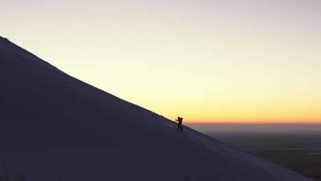 Ein-Freeskier-Klettert-Entlang-Der-Kante-Des-Berges-Zum-Gipfel,-Während-Im-Hintergrund-Der-Orangefarbene-Sonnenaufgang-Zu-Sehen-Ist