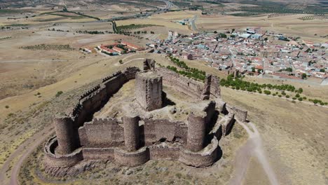 Orvital-flight-in-a-ruined-castle-with-its-walls-and-towers-on-a-hill-seeing-that-the-main-tower-is-in-the-middle-of-the-fortress-and-with-crop-fields-and-a-town-on-a-summer-day-in-Toledo-Spain