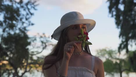 Hermosa-Mujer-Con-Sombrero-Despreocupado-Con-Olor-A-Rosa-Rodeada-De-Vegetación-Al-Atardecer