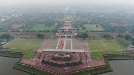 Drone-aerial-view-in-Vietnam-flying-over-Hue-imperial-stone-brick-fortress-wall,-green-gardens,-temples,-buildings-and-flag-on-a-cloudy-and-foggy-day