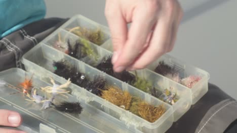 Compartmentalized-white-opaque-tackle-box-with-woven-feather-and-shimmering-flies-are-rearranged-by-angler