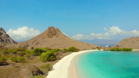 Vuelo-Sobre-La-Playa-Rosa,-Pantai-Merah,-En-Indonesia,-Capturando-Una-Impresionante-Perspectiva-Aérea-De-Su-Belleza-única-Y-Vibrante-De-Montañas-Escarpadas-Y-Agua-Turquesa-Con-Un-Cielo-Azul-De-Fondo.