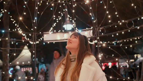 Beautiful-young-woman-with-dark-hair-and-white-Coat-smiling-straight-to-the-Camera-and-looking-up-to-the-Christmas-Lights-Decoration-on-a-Christmas-Market-during-a-winter-cold-evening-in-slow-motion