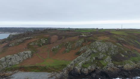Guernsey-Pleinmont,-flight-from-out-to-sea-towards-the-south-west-tip-of-the-Island-on-cloudy-day-showing-cliffs,-heathland,beach-rocks-with-foaming-waves-German-Watchtower-and-the-BBC-tower