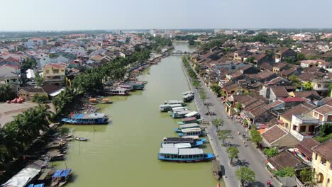 Drone-aerial-view-in-Vietnam-flying-over-Hoi-An-brown-color-river-canal-in-the-city,-small-brick-houses-and-wooden-boats-on-a-sunny-day