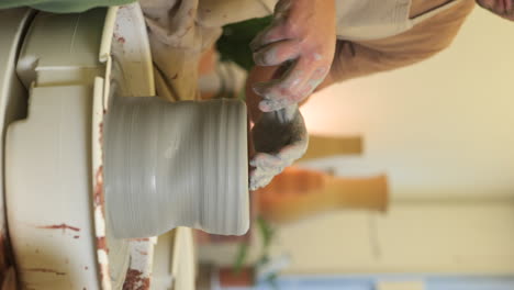 VERTICAL-male-potter-wearing-apron-sitting-at-workshop-turntable-shaping-clay-material