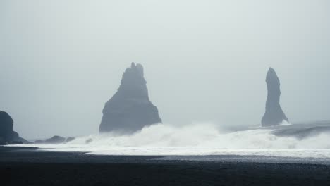 Tall-dark-basalt-towers-stand-out-amidst-a-foggy-black-sandy-beach-as-waves-crash-onto-the-shore