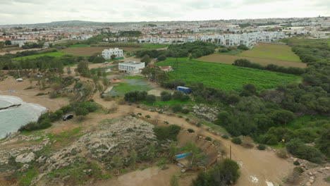 Aerial-view-of-a-coastal-area-blending-natural-and-urban-landscapes,-with-rocky-terrain-leading-to-agricultural-fields,-and-a-scattering-of-buildings-with-a-town-in-the-distance
