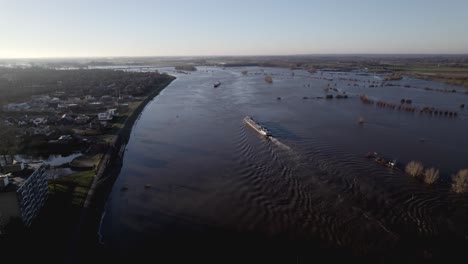 Aerial-approach-of-inland-shipping-large-cargo-vessels-leaving-ripple-waves-on-river-IJssel-during-high-water-level-with-flooded-floodplains-of-tower-town-Zutphen-in-The-Netherlands