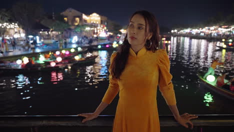 Young-Vietnamese-woman-posing-in-traditional-yellow-dress-at-night-river-scene-in-Hoi-An,-Vietnam